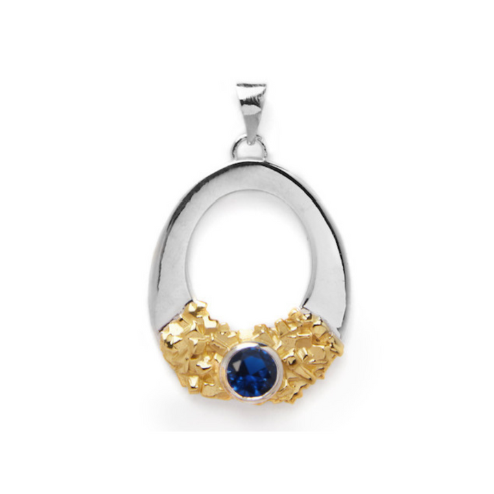 Introducing the Giselle Pendant by Gallardo & Blaine Designs: a stunning sterling silver oval pendant adorned with a cluster of small gold accents at the bottom, showcasing a large, round blue gemstone set elegantly in the center of the gold accents. The pendant features a simple silver bail at the top for easy attachment to a chain.