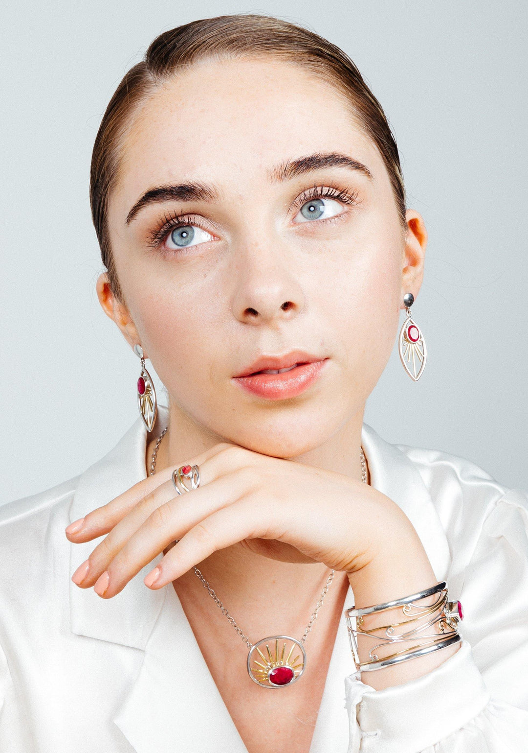 A young woman with light eyes and fair skin, wearing a white blouse, looks upwards. She showcases various sterling silver & gold jewelry pieces including the Goddess Earrings in various gemstones by Gallardo & Blaine Designs, a necklace with a red gem pendant, a ring with a red gem, and multiple bracelets. Her hair is pulled back neatly.