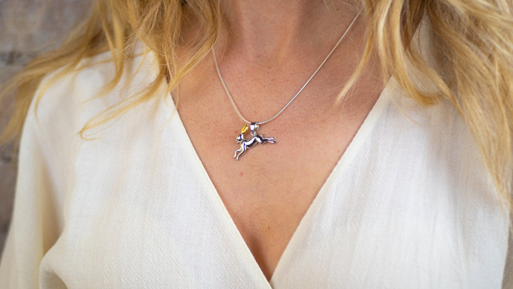 A close-up of a woman wearing a white V-neck top and a silver necklace from Gallardo & Blaine Designs, featuring an 18ct hard gold plating Wildlife Pendant - Hare With Chain. The focus is on the necklace and pendant, with her blonde hair partially visible in the background.