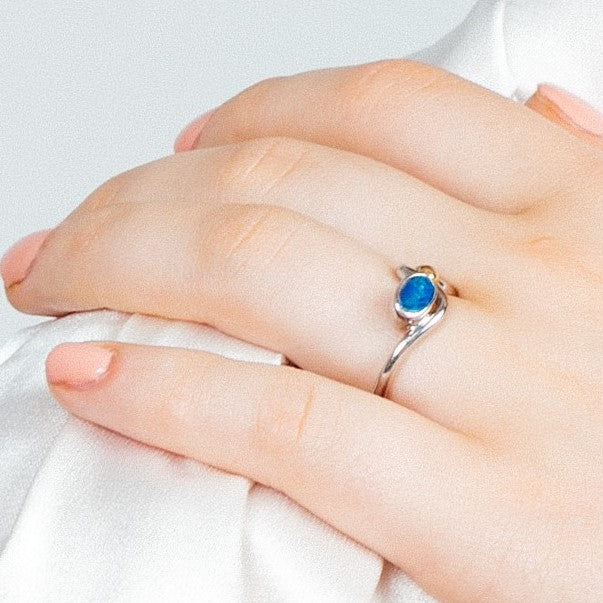 A close-up of a light-skinned hand wearing Gallardo & Blaine Designs' Adjustable Heartbeat Ring in sterling silver, featuring a small oval blue gemstone. The hand is on soft white fabric, with nails manicured and painted pale pink.