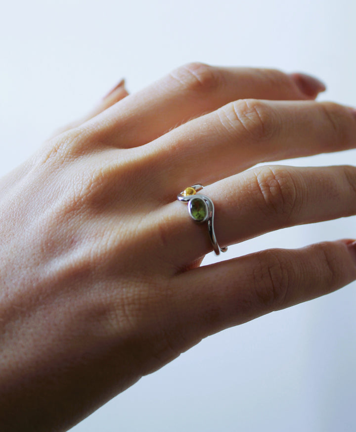 A manicured hand wears the Gallardo & Blaine Designs Adjustable Heartbeat Ring in sterling silver, with two gemstones—one light, one dark—set at each end of the open band, shown against a softly blurred background.