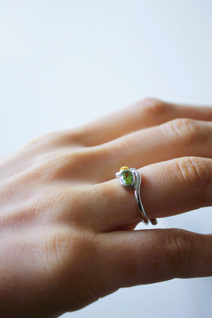 A close-up of a hand wearing Gallardo & Blaine Designs’ Adjustable Heartbeat Ring, crafted in sterling silver and featuring a small green gemstone, shown on the ring finger against a plain light background.