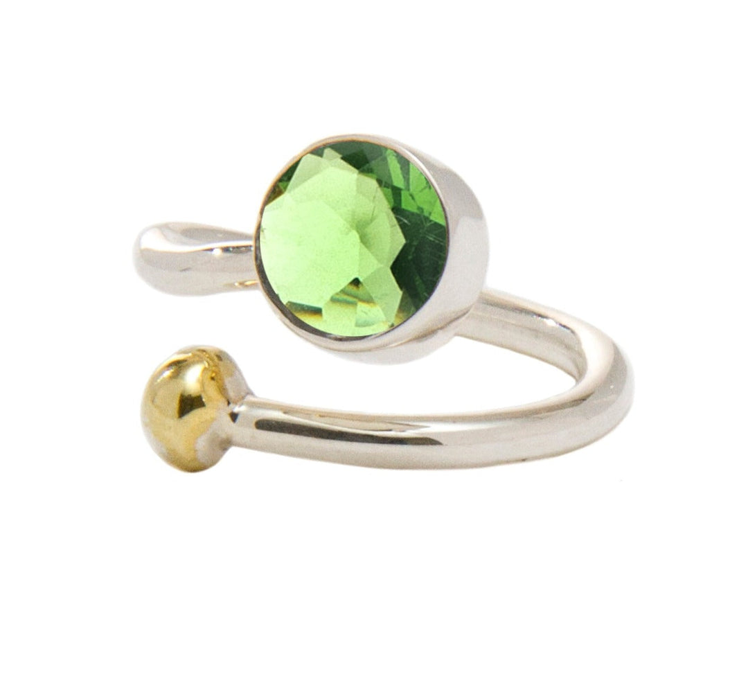 A shiny, open-ended silver and gold jewelry piece features a round, faceted green gem on one end and a small gold ball on the other. This elegant Honeysuckle Ring in Silver Gold & gemstones by Gallardo & Blaine Designs' minimalist design highlights the vibrant green stone and its contrasting gold accent.