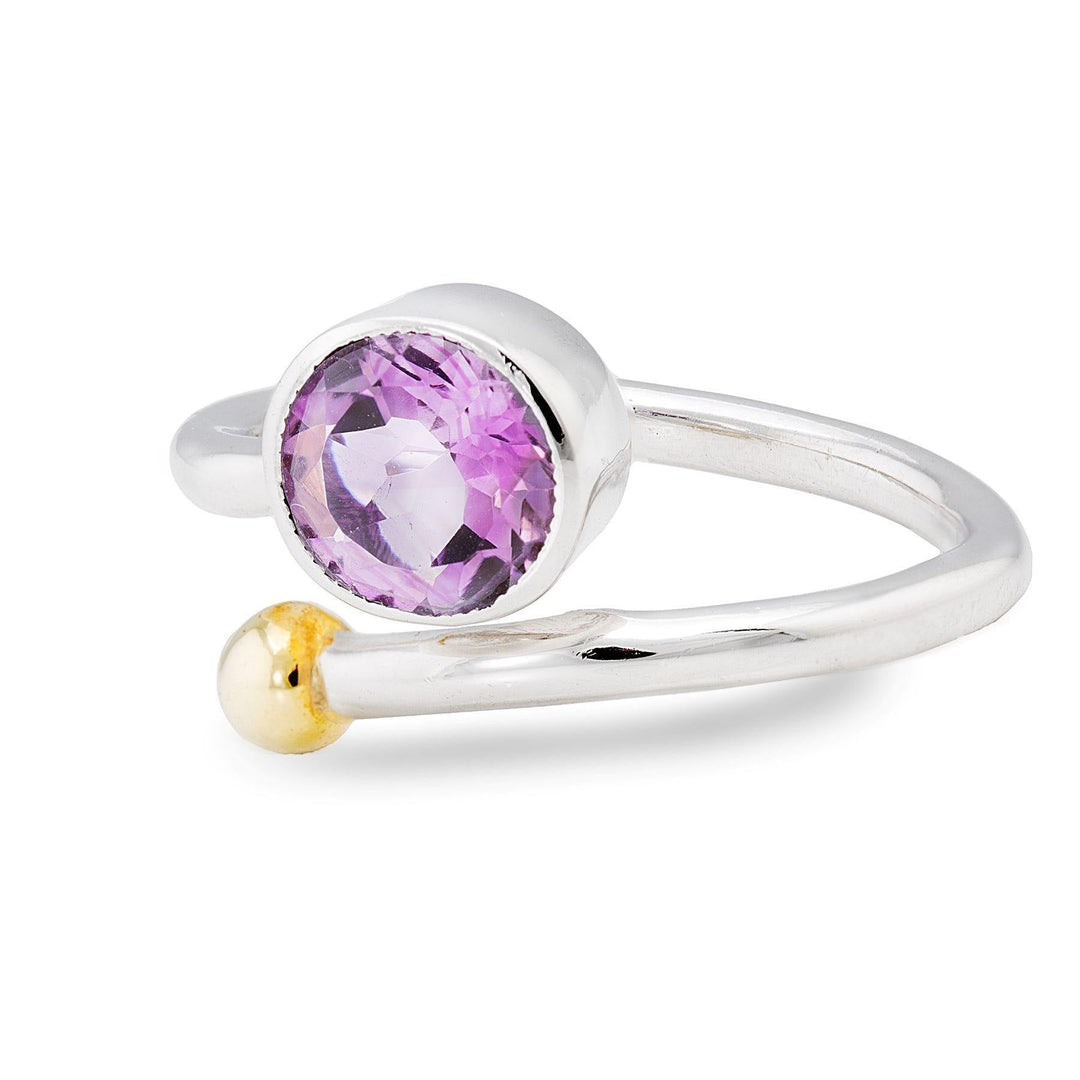 A close-up image of an elegant Honeysuckle Ring in Silver Gold & gemstones by Gallardo & Blaine Designs featuring a purple gemstone as its central focal point. The silver and gold jewelry piece has an open-ended band adorned with a small gold sphere at one end and the round-cut purple gemstone encased in silver at the other, reminiscent of a honeysuckle flower design.