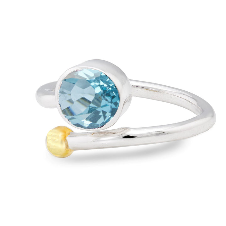 The Gallardo & Blaine Designs Honeysuckle Ring in Silver Gold & gemstones features an open band design with a blue, faceted gemstone set in a round bezel on one end and a small, gold-tone sphere on the other. This elegant adjustable gemstone ring captures a minimalist, modern aesthetic, perfect for lovers of silver and gold jewelry.