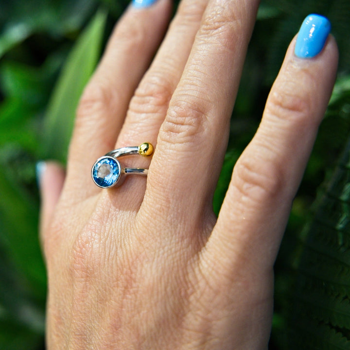 A hand with light blue nail polish wears Gallardo & Blaine Designs' Adjustable Honeysuckle Ring in Silver Gold & gemstones, showcasing a blue gemstone and small golden bead. The background is a blurred green, probably foliage.