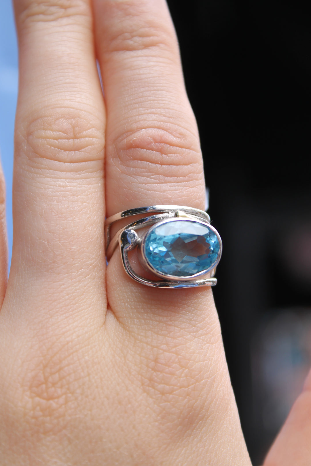 A close-up of a hand wearing Gallardo & Blaine Designs’ Horizontal Ring, featuring a large oval blue gemstone. The elegant sterling silver ring stands out against the blurred background.