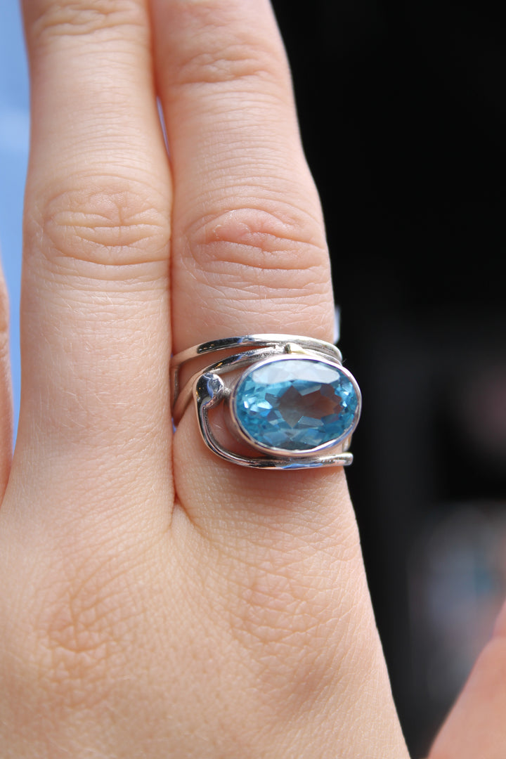 A close-up of a hand wearing Gallardo & Blaine Designs’ Horizontal Ring, featuring a large oval blue gemstone. The elegant sterling silver ring stands out against the blurred background.