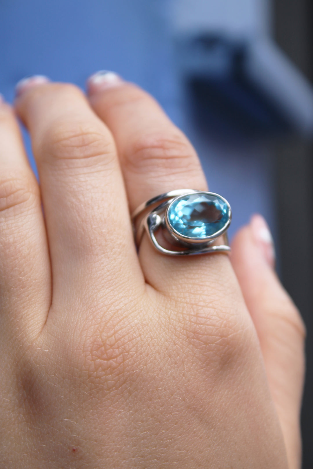 A manicured hand wears Gallardo & Blaine Designs’ Horizontal Ring, featuring a large oval blue gemstone, set in sterling silver against a blurred blue background.