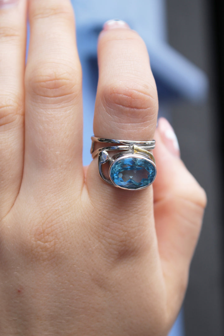 A close-up of a hand wearing Gallardo & Blaine Designs' Horizontal Ring, featuring a large oval blue gemstone, on the middle finger. The background is softly blurred in blue and black tones.