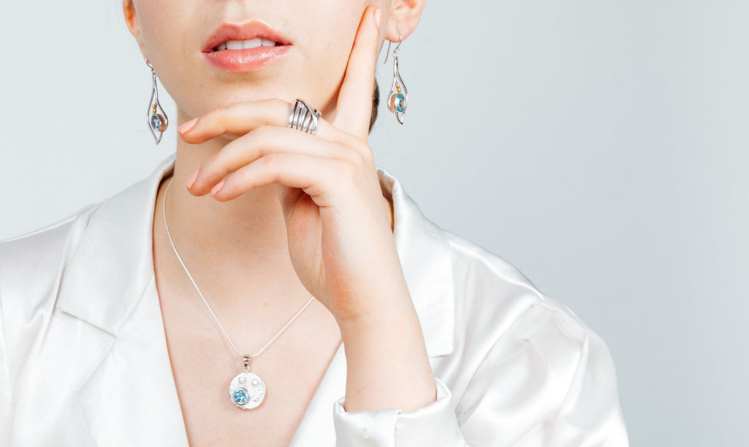 A person wearing elegant silver & gold jewelry, including Gallardo & Blaine Designs Iris Earrings in various gemstones, rings, and a necklace. The necklace features a round pendant with a blue gem. The person is dressed in a white satin blouse, with their hand gracefully touching their chin and lips slightly parted.