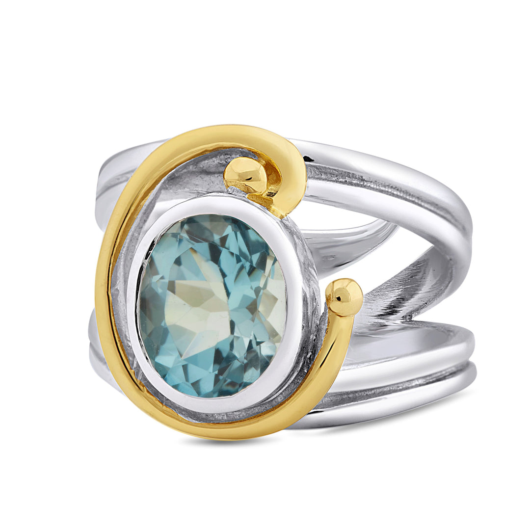 A silver ring with a split shank design features an oval blue gemstone encircled by a gold-tone spiral accent. This Jasmine Ring in various gemstones from Gallardo & Blaine Designs combines both silver and gold tones, giving it a unique and elegant appearance.