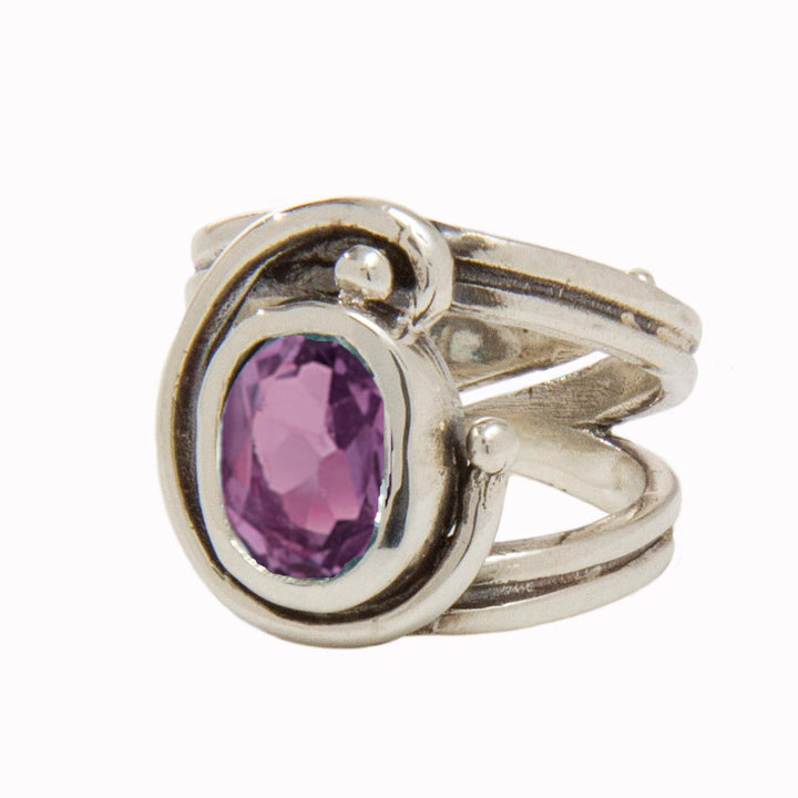 A silver Jasmine Ring in various gemstones by Gallardo & Blaine Designs features an oval-shaped, faceted purple gemstone set in the center, surrounded by an intricate swirl design. The thick silver band is split into three parts where the swirl design attaches, adding to its adjustable design.