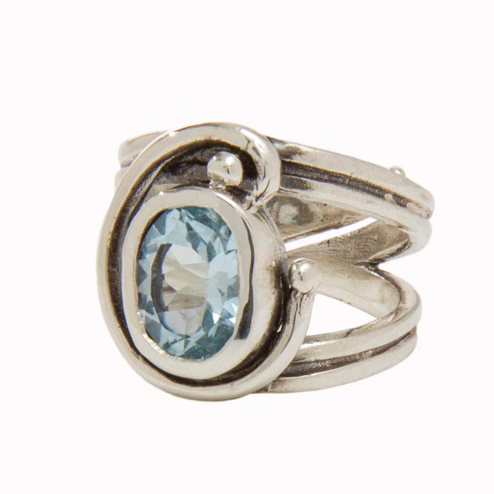 A Jasmine Ring in various gemstones by Gallardo & Blaine Designs features a large, oval-shaped blue gemstone set in the center of an intricate, swirling band design. The ring's adjustable design offers a modern and elegant appearance.