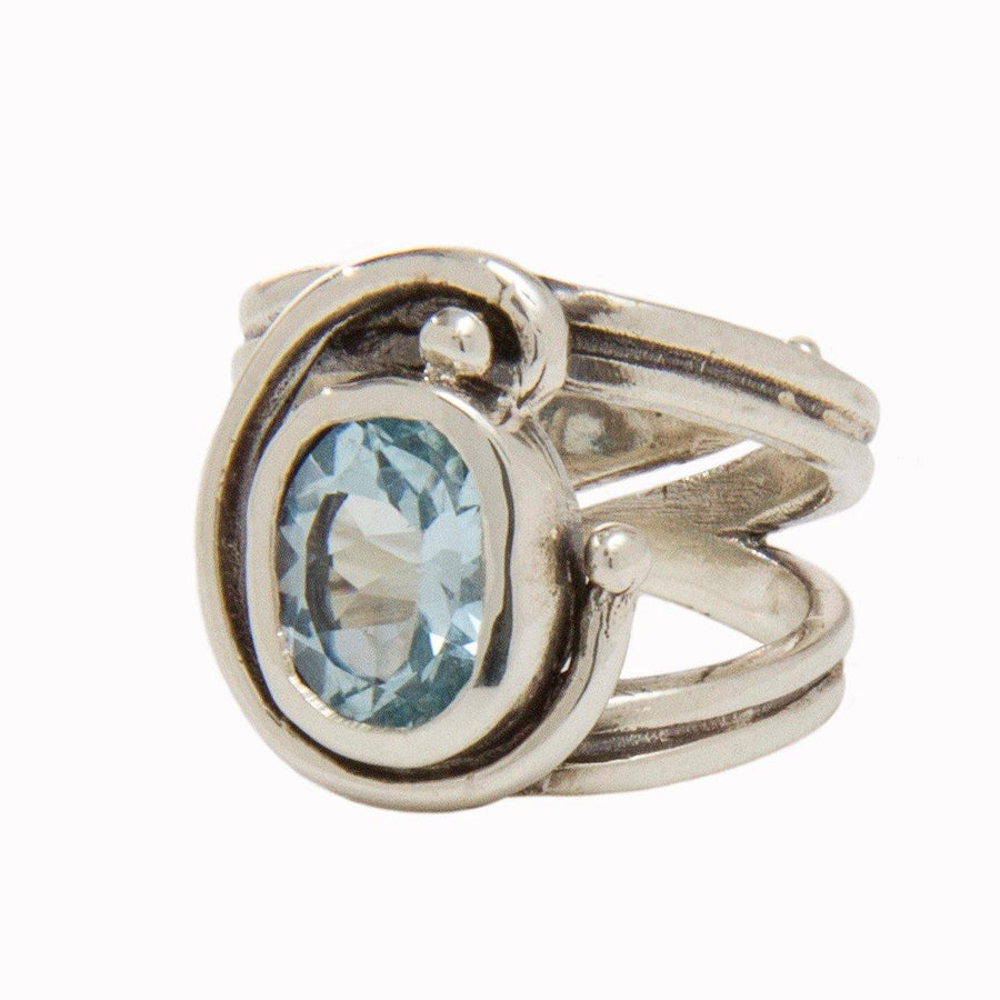 A Jasmine Ring in various gemstones by Gallardo & Blaine Designs features a large, oval-shaped blue gemstone set in the center of an intricate, swirling band design. The ring's adjustable design offers a modern and elegant appearance.