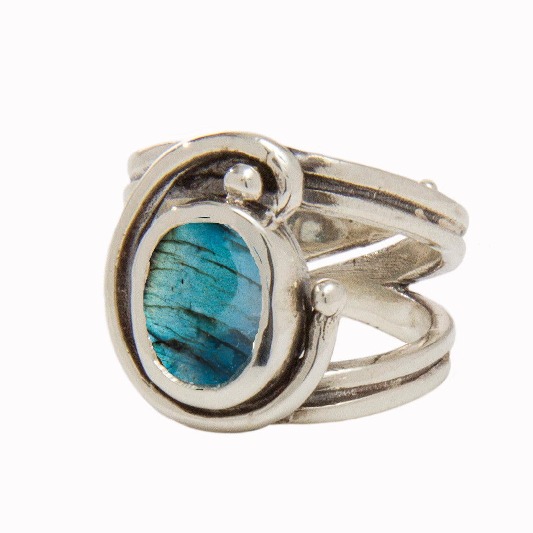 The Jasmine Ring in various gemstones by Gallardo & Blaine Designs features an intricate design with a prominent blue-green oval gemstone at the center. The band splits into decorative swirls encircling the gemstone, showcasing a unique and artistic style with an adjustable design for comfort and elegance.