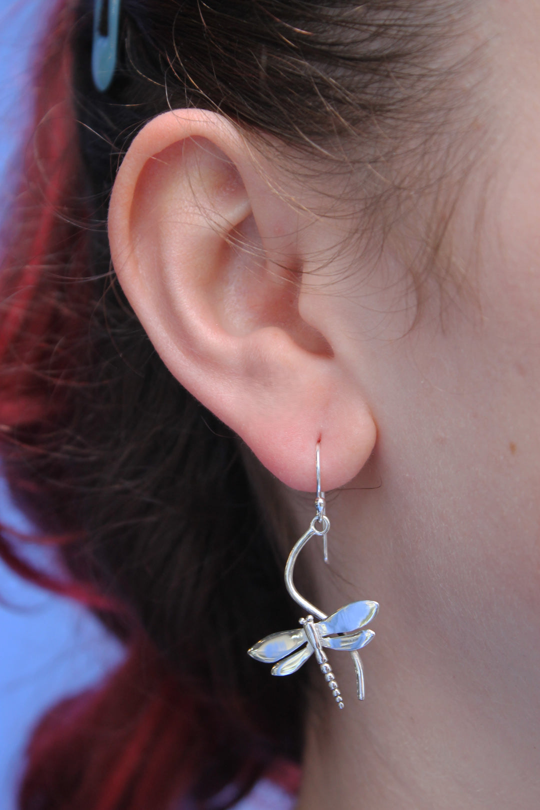 A close-up shows a person with reddish hair, pulled back, wearing Gallardo & Blaine Designs' Wildlife Juniper Earrings (Silver)—dragonfly-shaped jewelry—against a light blue background.