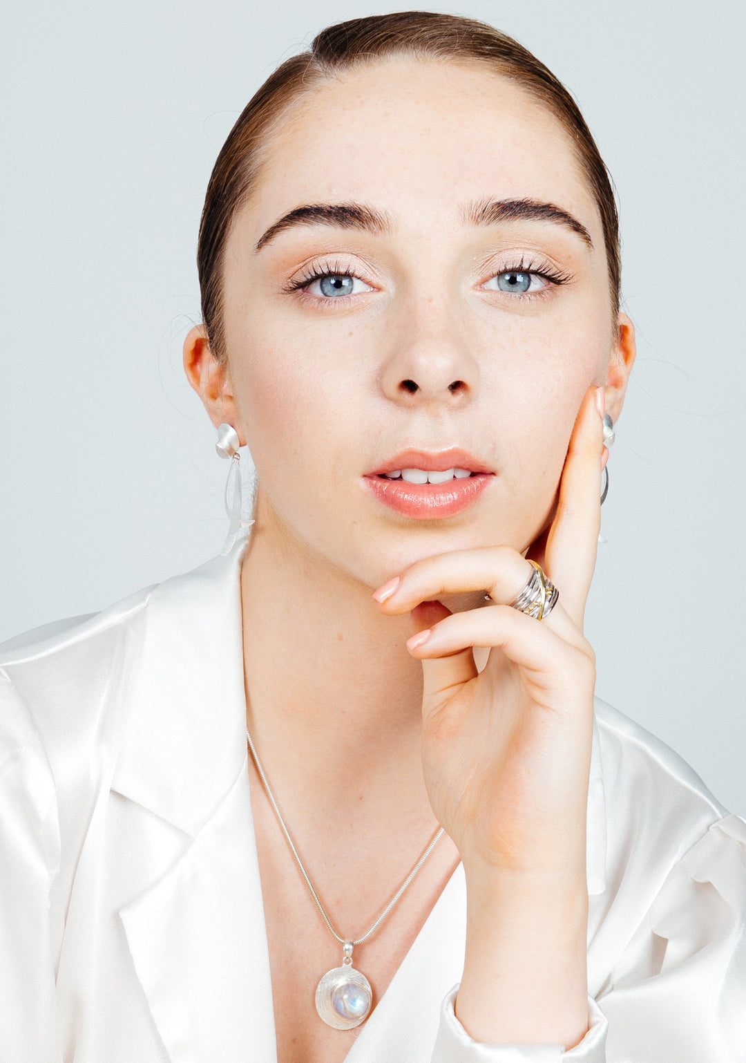 A person with light skin, blue eyes, and brown hair pulled back, is wearing minimal makeup, a white satin top, Juno Earrings in various gemstones by Gallardo & Blaine Designs, and a necklace with a pearl pendant. They rest their chin on their hand, displaying a calm and thoughtful expression.