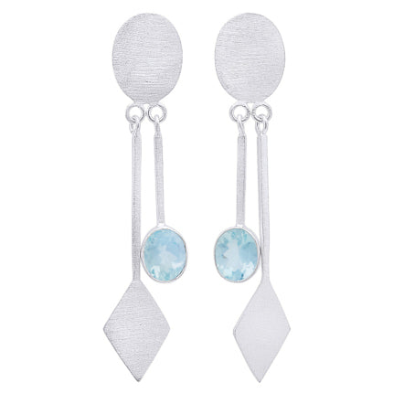 A pair of modern fashion jewelry, the Karo Earrings in various gemstones by Gallardo & Blaine Designs feature brushed sterling silver ovals at the top. Each earring has a long, narrow bar dangling from the oval, ending in a diamond shape and a light blue gemstone midway down the bar.