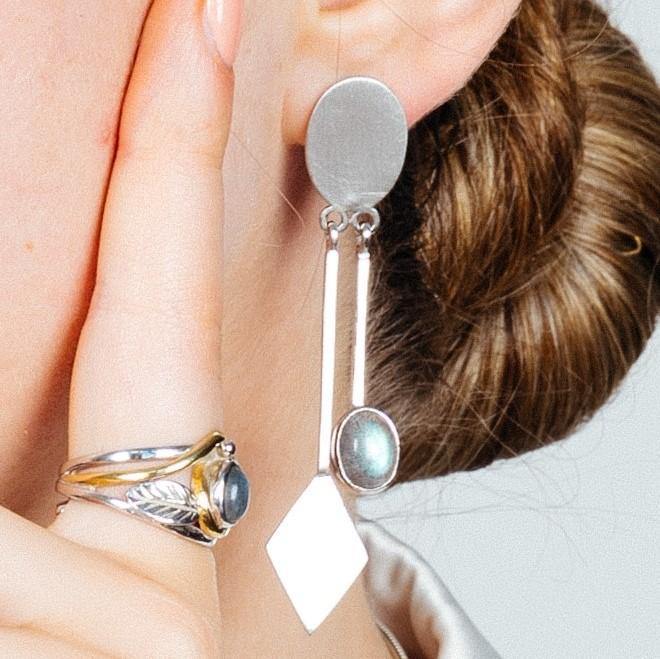 Close-up of a person's ear wearing the elegant Karo Earrings in various gemstones by Gallardo & Blaine Designs, featuring oval and diamond-shaped elements with a reflective surface. The person also wears a silver ring with a large, round, greyish-blue stone, and their hair is styled in a low bun, epitomizing modern fashion jewelry.