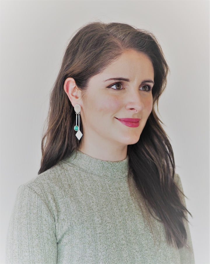 A woman with long, wavy brown hair is smiling softly. She is wearing a light green sweater and Karo Earrings in various gemstones by Gallardo & Blaine Designs that dangle elegantly. The background is plain and muted, making her the focal point of the image.