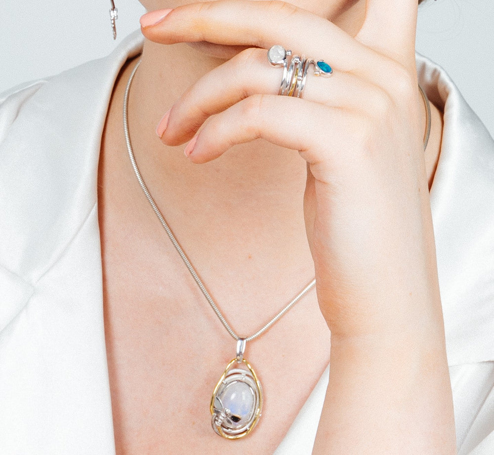 A person wearing a white blouse showcases a Lagoon Pendant Large in various gemstones by Gallardo & Blaine Designs, a silver necklace with an intricately designed dragonfly featuring a central white gemstone. The person also wears multiple rings, including one with a blue stone and others with various designs.