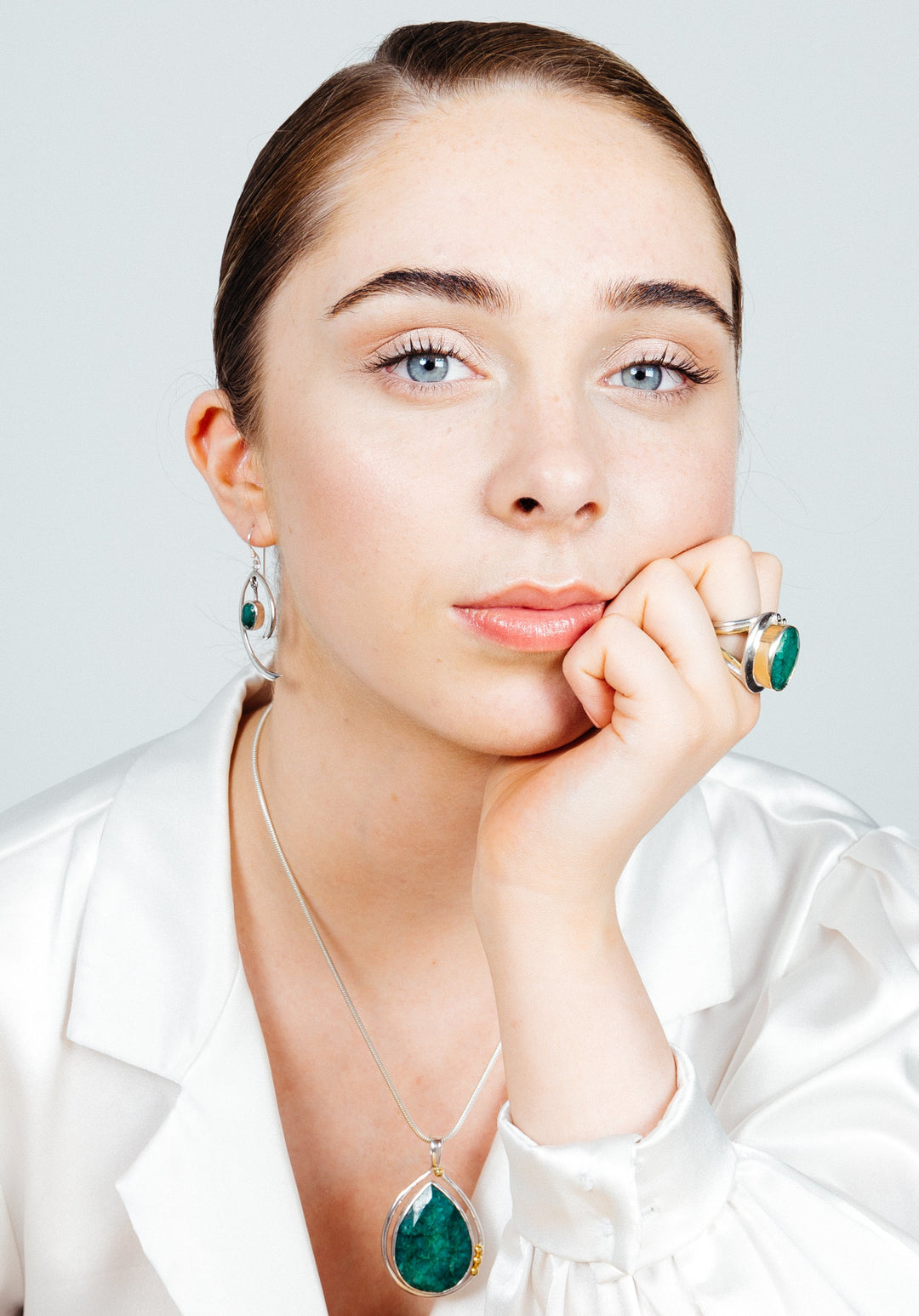 A woman with light skin and blue eyes gazes directly into the camera, resting her chin on her hand. She exudes minimalist elegance in a white blouse and matching green gemstone jewelry, including a necklace, Primrose Earrings in various gemstones from Gallardo & Blaine Designs, and rings. Her hair is pulled back neatly.