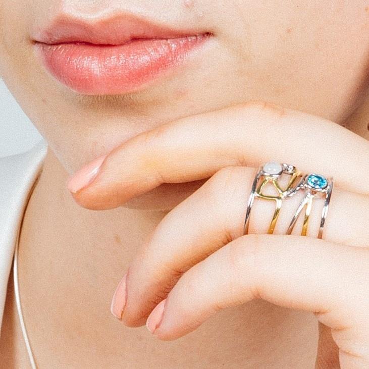 Close-up of a person’s face and hand. The person is wearing a Gallardo & Blaine Designs Lattice Ring in various gemstones—blue, clear, and gold accents—on their index finger. Their lips are softly closed, and the hand adorned with contemporary design touches their chin gently.