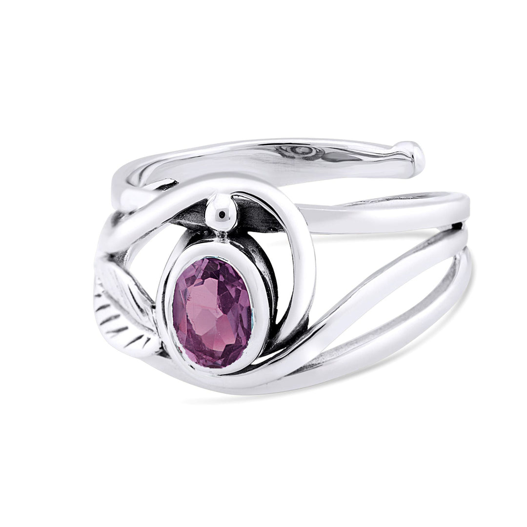 A silver and gold ring features an intricate, twisting design with a central, oval-shaped purple gemstone. The band splits into two delicate loops on each side of the gemstone, with a small leaf detail on one end, adding an elegant touch. This Leaf Ring in Silver Gold & various gemstones by Gallardo & Blaine Designs is truly captivating.