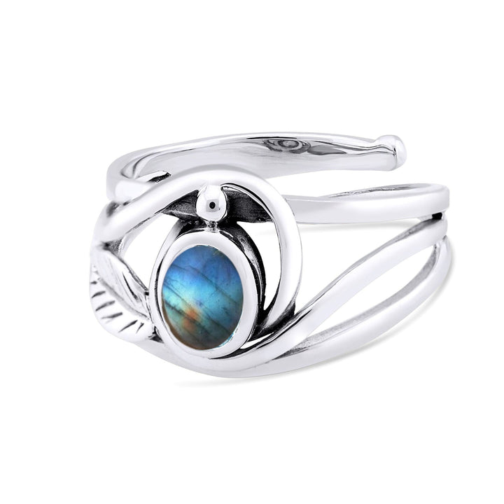 The Leaf Ring in Silver Gold & various gemstones by Gallardo & Blaine Designs features a unique, intertwined band design, with an oval-shaped labradorite gemstone set in the center. The gemstone displays a range of iridescent colors, mainly blue and green. This elegant gemstone ring has an artistic look that captivates the eye.