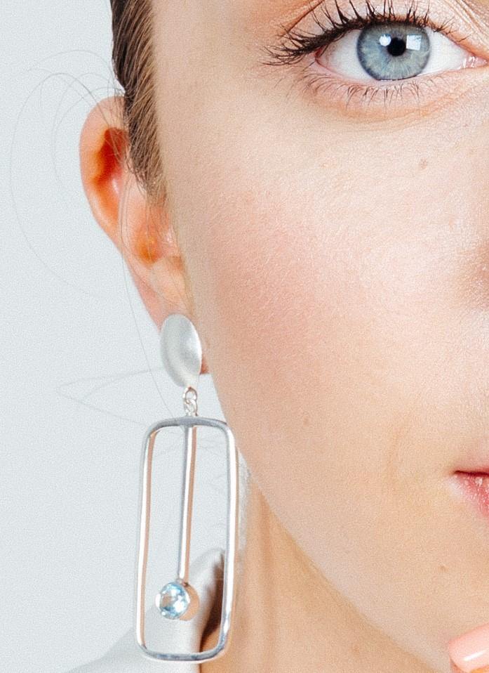 Close-up of the right side of a person’s face, focusing on a blue eye and a unique Gallardo & Blaine Designs Lorelei Earrings Large in various gemstones. The geometric silver earring consists of an elongated oval shape with an attached circular element and a small blue gemstone. The image is well-lit, highlighting skin texture and details.