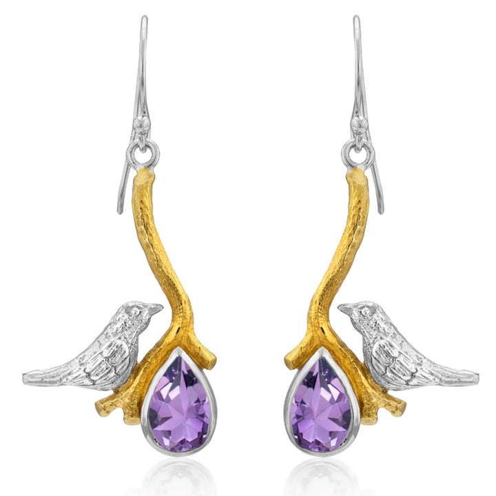 A pair of elegant Lovebird Earrings in various gemstones from Gallardo & Blaine Designs featuring a design with silver birds perched on gold branches. Each branch holds a teardrop-shaped purple gemstone, adding a touch of color and sophistication to these statement earrings.