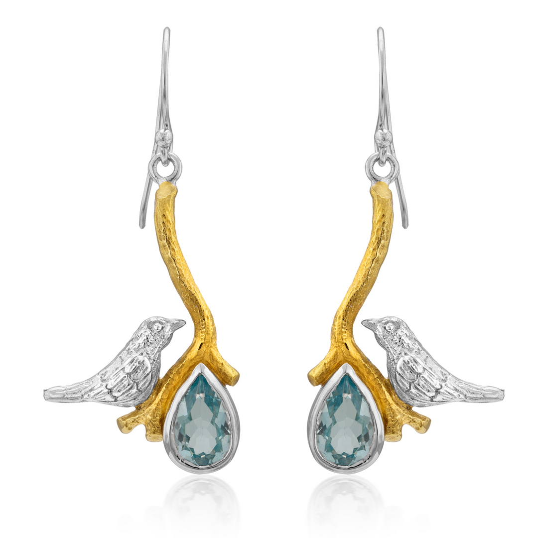 A pair of Lovebird Earrings in various gemstones by Gallardo & Blaine Designs featuring silver birds perched on gold-tone branches. Below each bird hangs a teardrop-shaped blue gemstone, creating elegant dangle drop earrings. The branches connect to silver hooks for wearing.