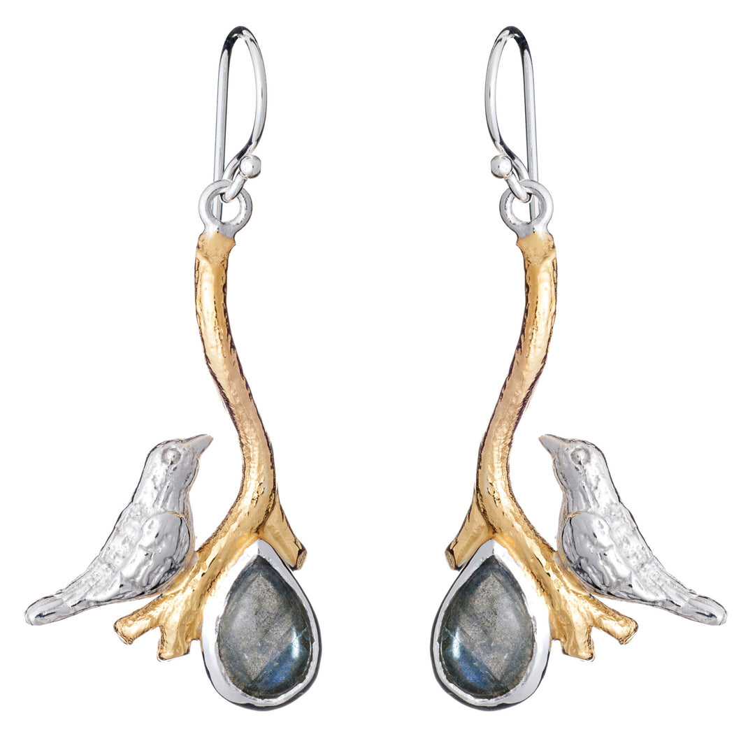 A pair of Lovebird Earrings in various gemstones featuring silver birds perched on golden branches. Below each bird hangs a teardrop-shaped pendant with a dark, reflective gemstone. These Gallardo & Blaine Designs dangle drop earrings have hook-style backings for wearing.