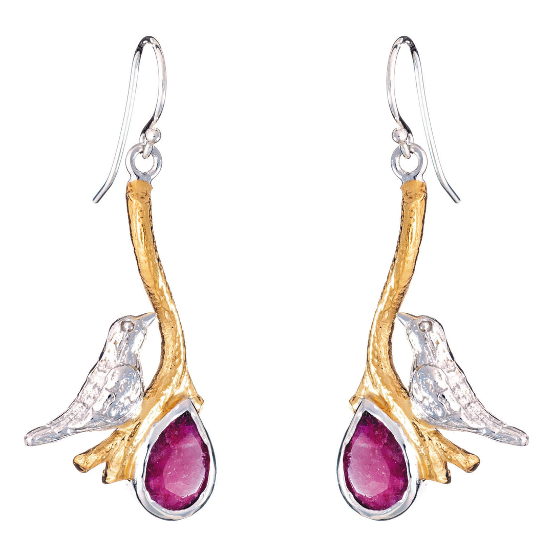 A pair of Lovebird Earrings in various gemstones from Gallardo & Blaine Designs features golden branches with silver birds perched on them, holding drop-shaped, deep red gemstones. The birds and branches are detailed with a shiny silver finish, while the gold adds a touch of elegance.