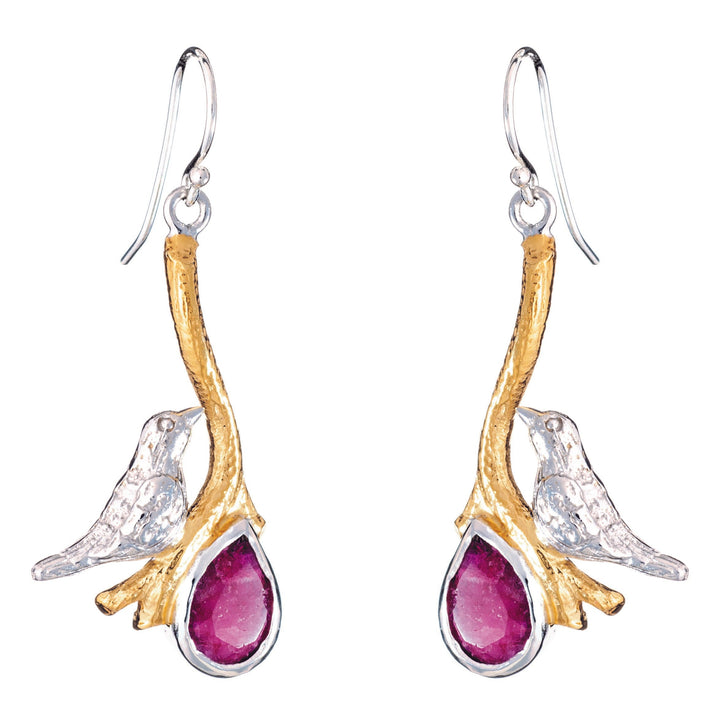 A pair of Lovebird Earrings in various gemstones from Gallardo & Blaine Designs features golden branches with silver birds perched on them, holding drop-shaped, deep red gemstones. The birds and branches are detailed with a shiny silver finish, while the gold adds a touch of elegance.