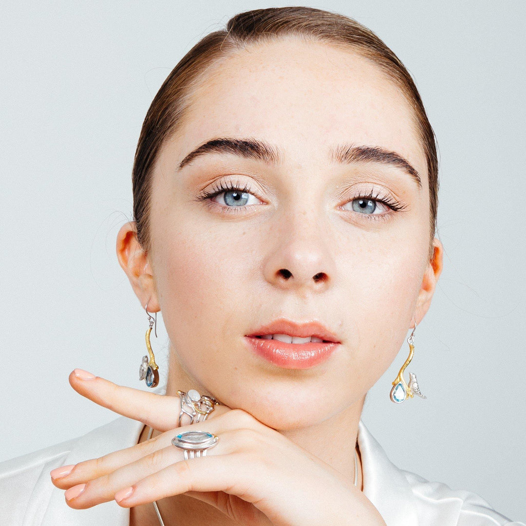 A young woman with fair skin and blue eyes gazes into the camera. She wears Gallardo & Blaine Designs' Lovebird Earrings in various gemstones and a ring with a large stone on her finger, which is gently resting near her chin. She has light makeup and her hair is pulled back. She wears a white top.