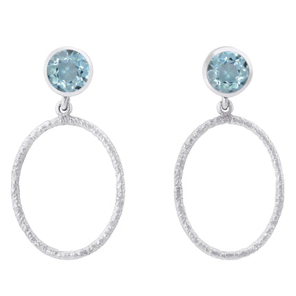 A pair of Gallardo & Blaine Designs Lunar Earrings in various gemstones featuring a small round blue gem at the top, attached to a large textured open circular hoop. The design is simple yet elegant, with the blue gems adding a touch of color to the sleek silver hoops.