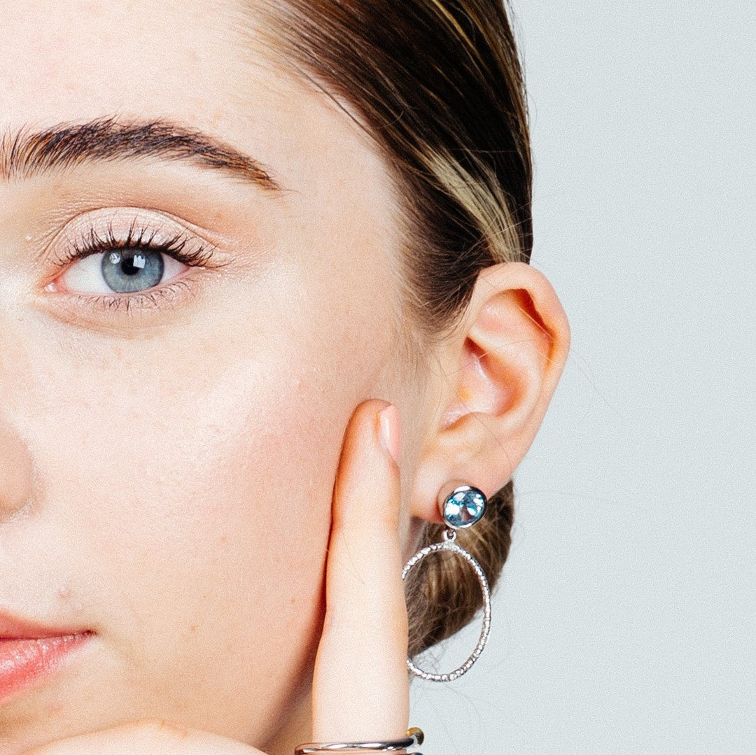 A close-up of a woman touching her earlobe with her index finger. She is wearing Lunar Earrings in various gemstones by Gallardo & Blaine Designs with a turquoise gemstone and a circular hoop. The image focuses on her ear and part of her face, showcasing smooth skin and light makeup.