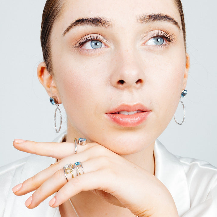 A person with light skin and short dark hair is looking upward, resting their chin on their hand. They are wearing Lunar Earrings in various gemstones by Gallardo & Blaine Designs adorned with dark beads and multiple silver rings. They have a thoughtful expression and are dressed in a white top.