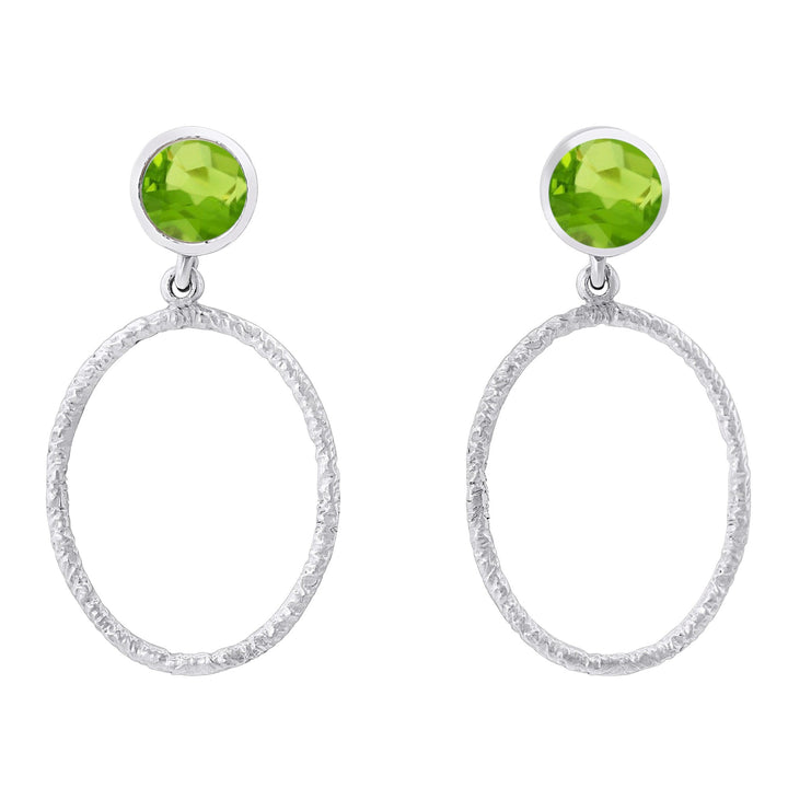 These Lunar Earrings in various gemstones by Gallardo & Blaine Designs feature a bright green gemstone at the top and a textured oval silver hoop dangling below. The design is elegant, with the green gemstone adding a pop of color to the minimalist silver hoop.