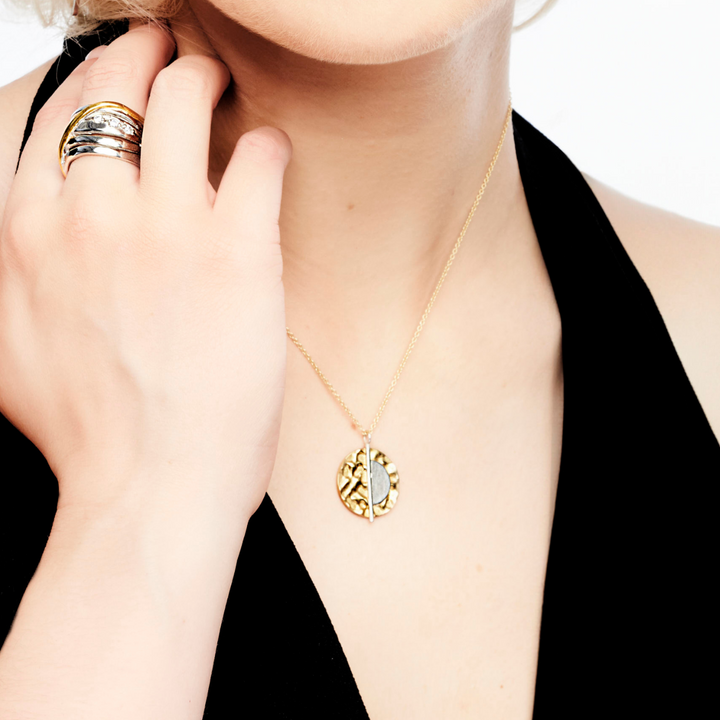 A person with light skin wearing a black sleeveless top holds their chin with their left hand, showcasing a sterling silver and gold vermeil ring on their index finger. The person also wears Gallardo & Blaine Designs' Lunar Eclipse Pendant & Chain in gold, featuring a round, intricate pendant. The background is plain white.