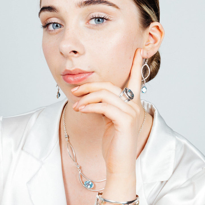 A woman with fair skin, blue eyes, and light brown hair is wearing a white satin blouse. She has on matching silver jewelry, including a necklace, rings, and a bracelet, all featuring blue gemstones. Her elegant drop silver earrings from Gallardo & Blaine Designs' Lupin Earrings in various gemstones collection glint softly as she rests her chin thoughtfully on her hand.