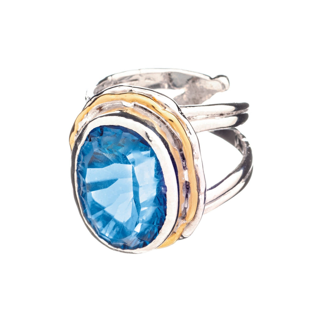 The Magnolia Ring in silver gold & various gemstones by Gallardo & Blaine Designs is a stunning statement piece crafted from sterling silver. It features a large, faceted blue gemstone as its oval-shaped centerpiece, accented by gold-toned bands that enhance its ornate design. With intricate detailing and an adjustable band, this ring exudes elegance.