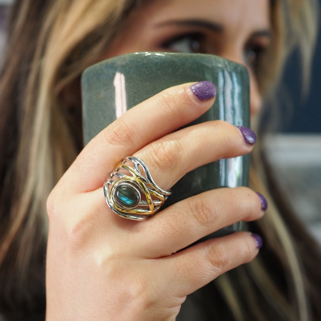 Close-up of a person holding a green ceramic mug with their right hand. They are wearing a large, intricate silver and gold chunky statement ring featuring a blue-green stone from the **Mayhem Ring in various gemstones by Gallardo & Blaine Designs**. The person's nails are painted purple with glitter. The background is blurred, focusing on the hand and mug.