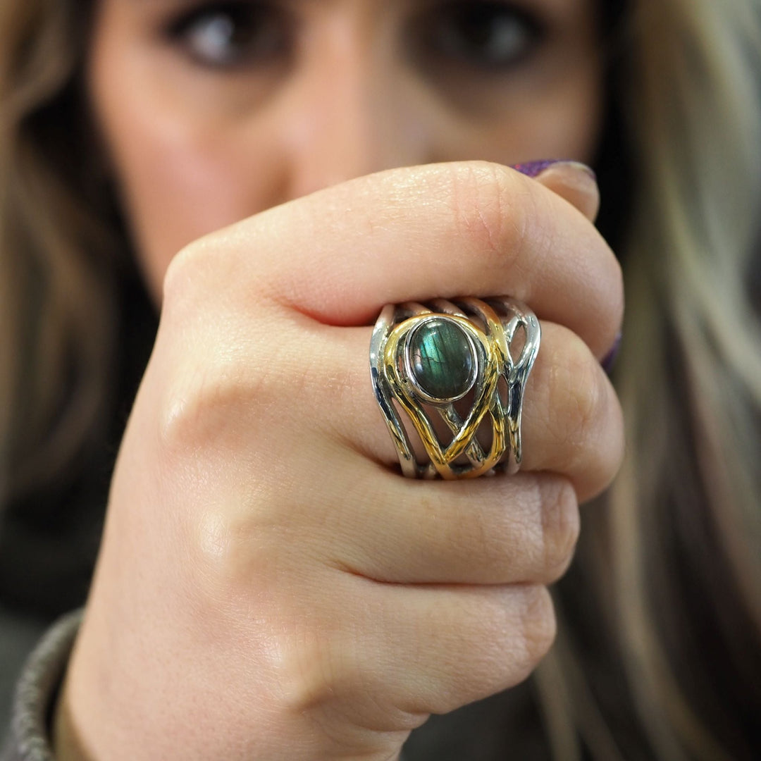 A close-up image of a person showing their hand with a striking, medieval-inspired chunky statement ring. The Mayhem Ring in various gemstones by Gallardo & Blaine Designs has an ornate design with intertwined silver and gold bands, featuring a large, round dark green stone. The out-of-focus face of the person is in the background.