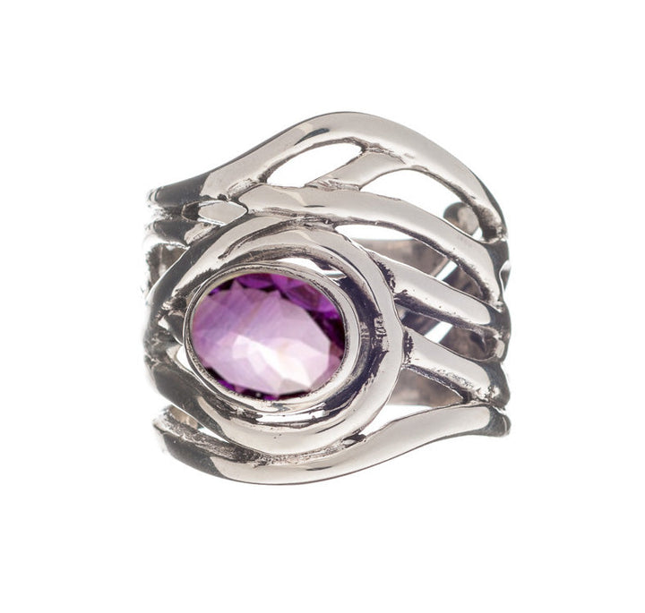 A Gallardo & Blaine Designs Mayhem Ring in various gemstones, featuring an intricate, wavy band design with a large oval-shaped purple gemstone set in the center. The chunky statement ring has a faceted gemstone that adds a reflective quality to the piece, giving it a modern and artistic yet medieval-inspired jewelry appearance.