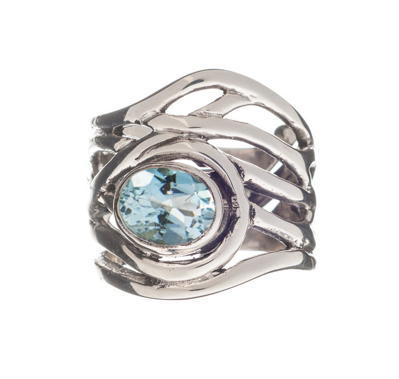 The Mayhem Ring in various gemstones from Gallardo & Blaine Designs features an intricate, wavy design and a large, round blue gemstone set in the center. The stone is faceted, catching the light and adding sparkle to this chunky statement ring reminiscent of medieval-inspired jewelry.