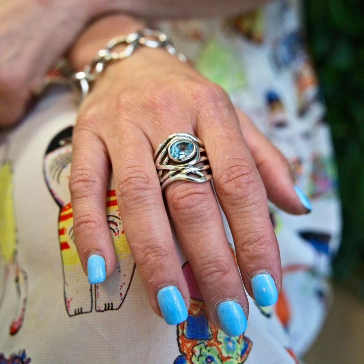 Close-up of a hand with blue painted nails, wearing the Mayhem Ring in various gemstones by Gallardo & Blaine Designs. The person is also adorned with a silver bracelet. The background features a colorful patterned fabric with whimsical designs.