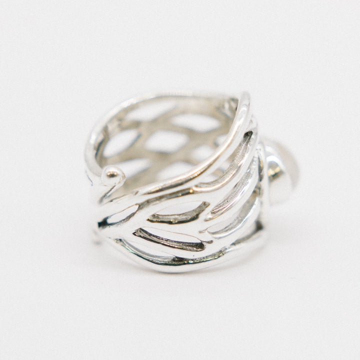 A close-up image of a chunky statement ring with an intricate, openwork design. The band features a pattern resembling interwoven strands, giving it a light and airy appearance. The Mayhem Ring in various gemstones by Gallardo & Blaine Designs is positioned slightly tilted, showcasing the detailed craftsmanship.